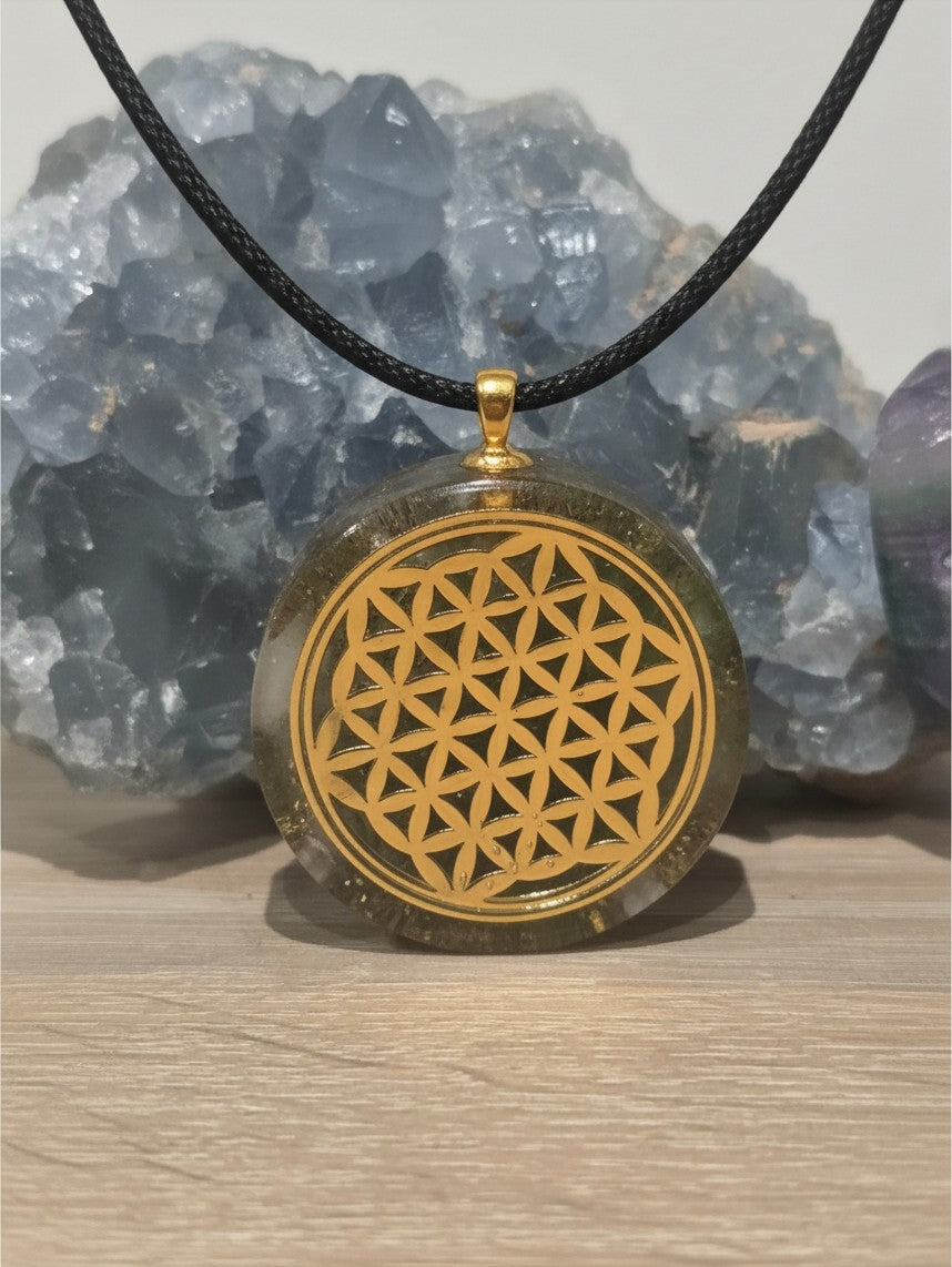 Pendentif orgonite AlyJoyCreation rond avec symbole doré Fleur de Vie sur cordon noir devant un cristal bleu